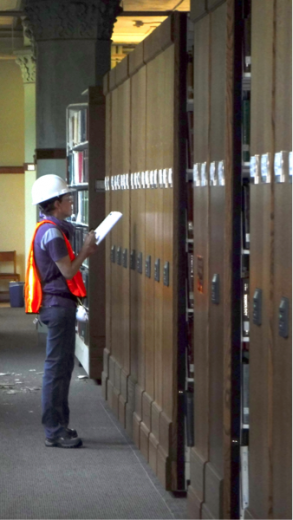A women in an orange vest and a white hard hat is inspecting the brown compact shelves.