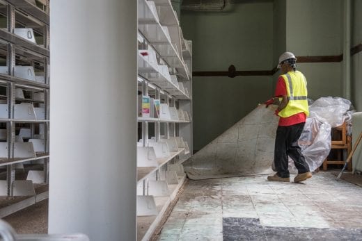 A Belfor worker in a yellow vests lifts up carpet next to white bookshelves. 