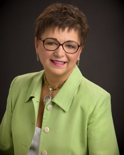 A headshot of a women with short brown hair, brown round glasses and a green button up shirt. 