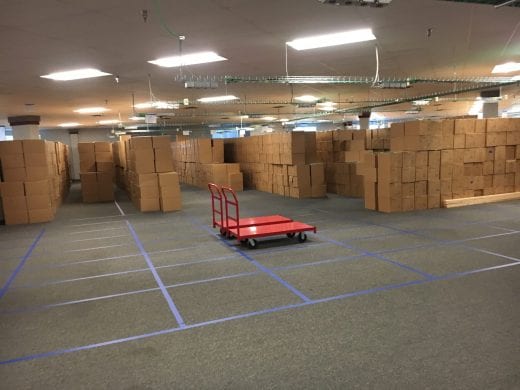 Blue tape lines the floor in grids and brown boxes are stacked in the boxes. 