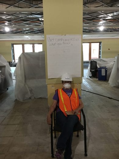 A woman in an orange vest and a white hard hat sits in a chair under a sign that reads get comfy and tell us what you think. 