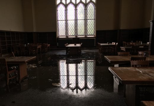 In a dimly lit room, the reflection of large leaded glass windows is visible in water pooled on the carpet. 