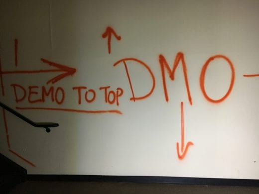 Orange spray paint on a yellow wall reads "Demo to top" and "DMO"