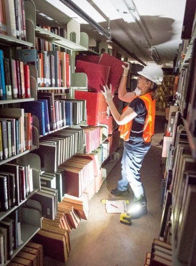 Woman wearing white hard hat, ventilation mask and neon orange emergency vest works by flashlight and pulls a large red book off of a bookshelf. 
