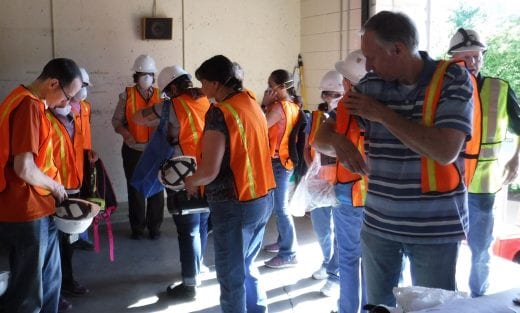 A group of twelve people dons orange emergency vests, hard hats and respirators. 