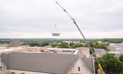 The metal arm of the crane extends high above the tan, peaked roof of the library as it lifts a large, rectangular piece of the HVAC unit.