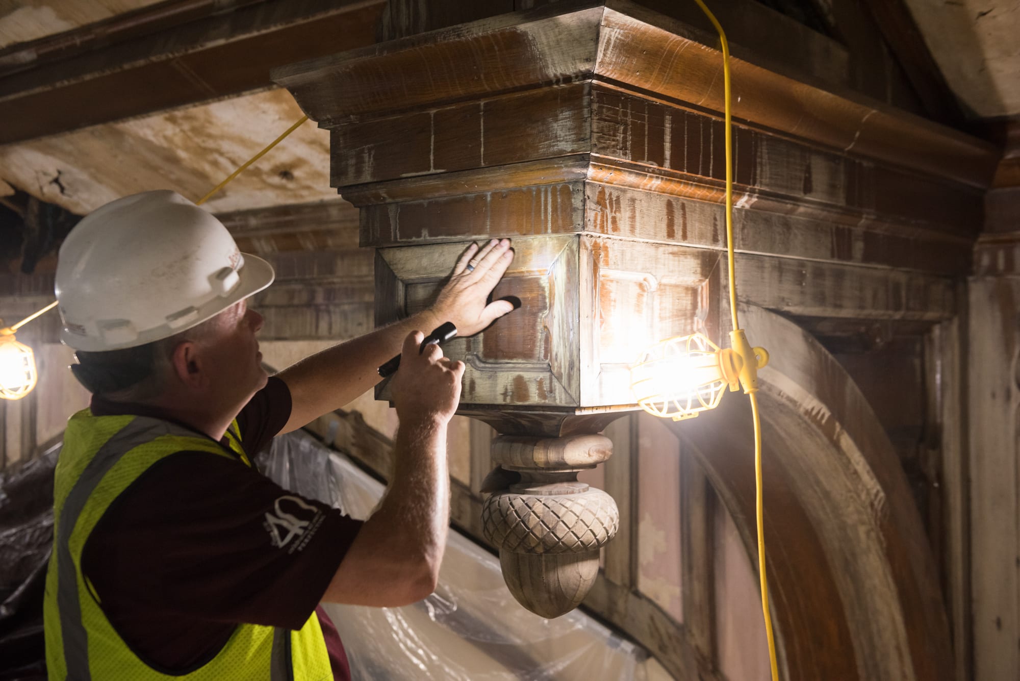 A man in a white hardhat and yellow construction vest faces a wood beam and examines it with a flashlight. 