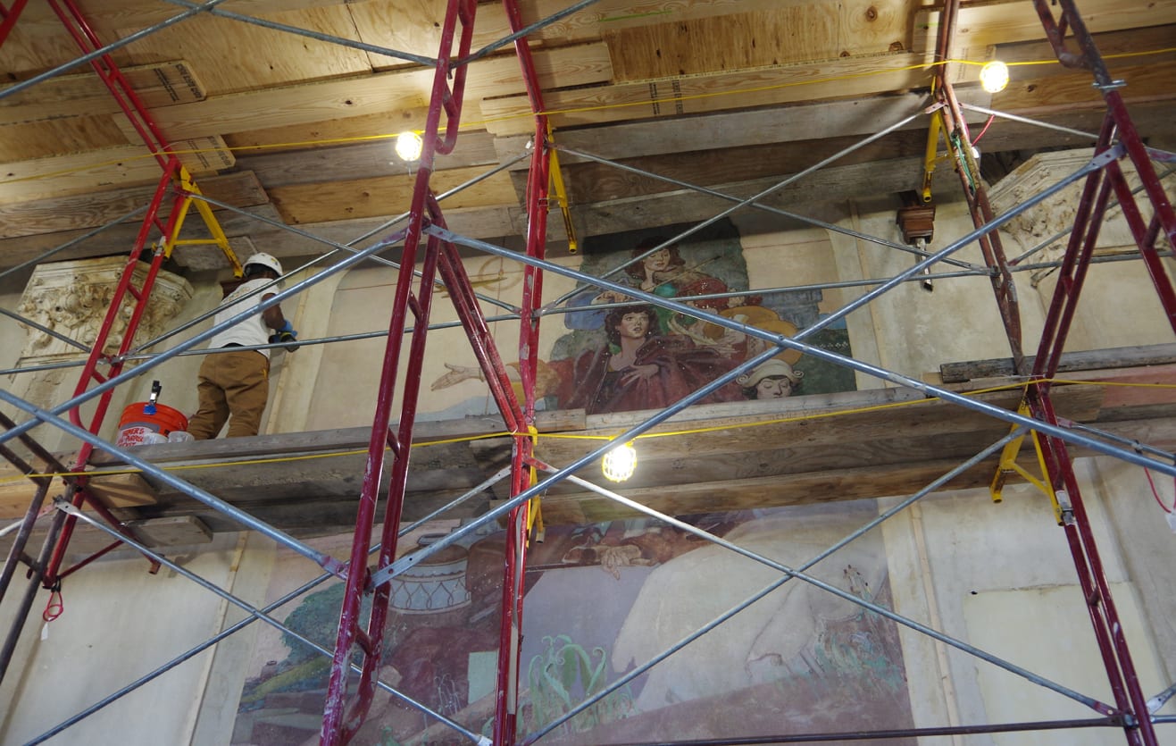 At left, a worker in a white hard hat stands elevated on a rough wood floor supported by scaffolding. An allegorical mural depicting the arts is to his right. 