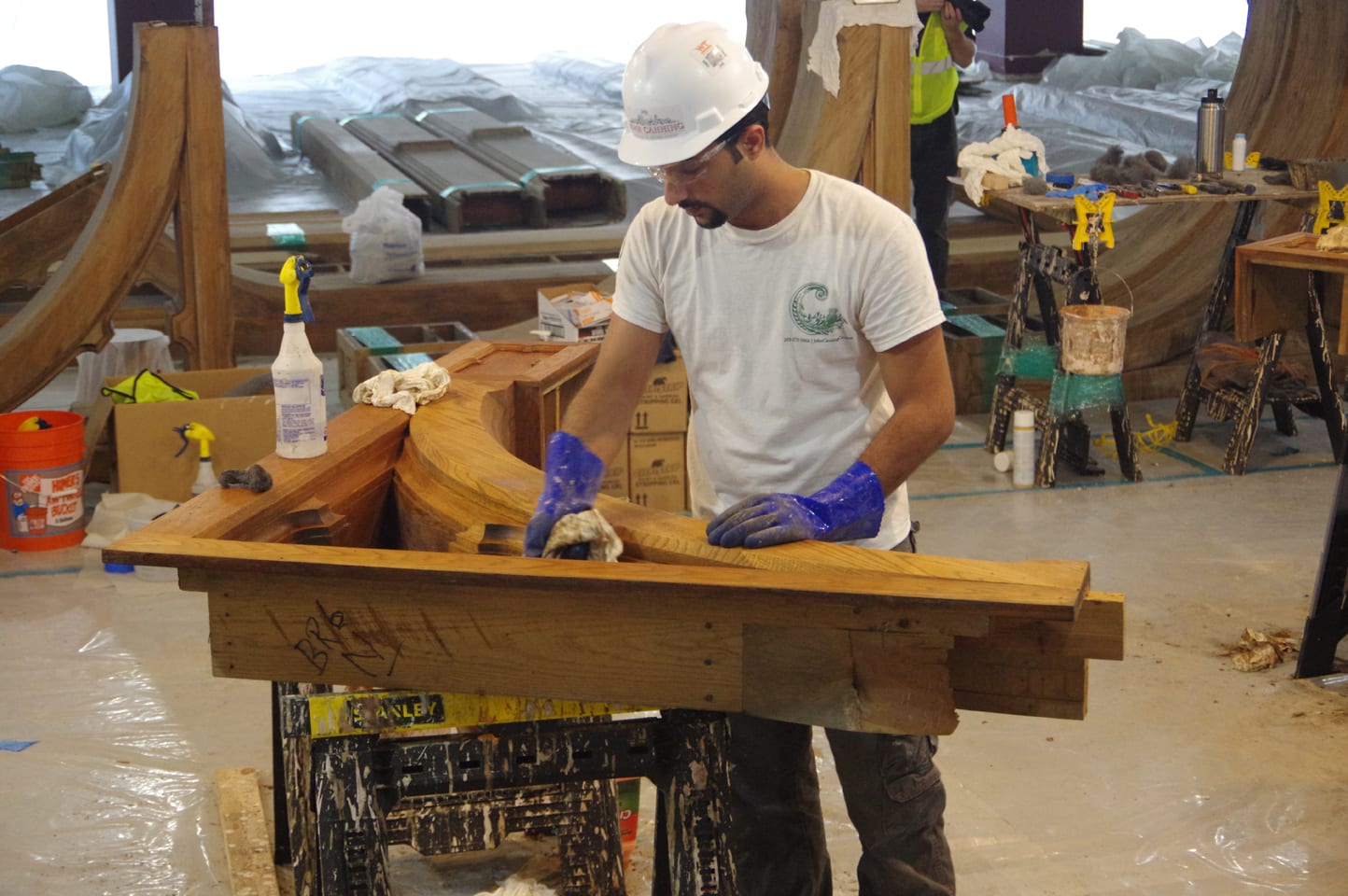 A dark-haired man in a white t-shirt and hardhat uses a cloth to wipe a heavy piece of wood sitting on sawhorses. 