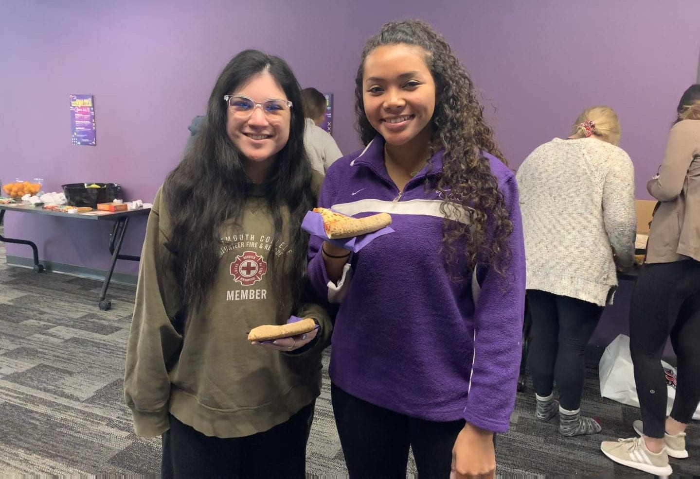 Two students smile holding pizza. 