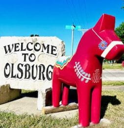 Red painted dala horse in front of Welcome to Olsburg sign