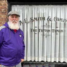 Man with gray beard and wearing hat standing in front of business window, Tipton Times